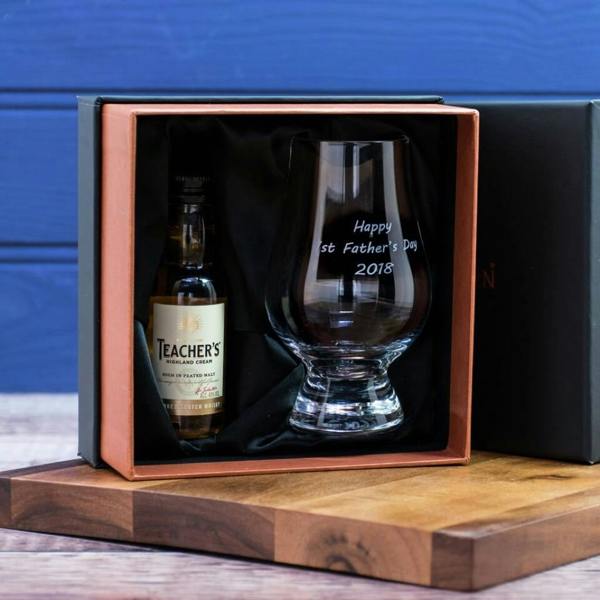 A black and rose-gold presentation gift box, open to reveal a personalised whisky glass etched with the text "Happy 1st Father's Day 2018" alongside a miniature bottle of Teacher's Highland Cream whisky, resting on a wooden board.