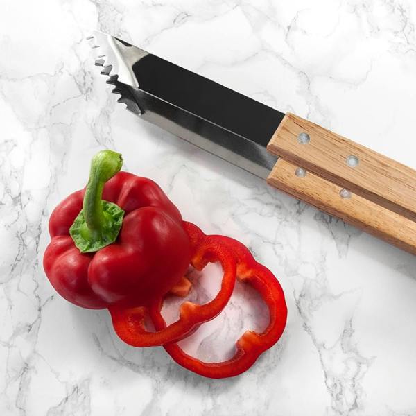 A close-up view of heavy-duty stainless steel barbecue tongs with natural wooden handles, styled next to a sliced fresh red bell pepper on a white marble background.
