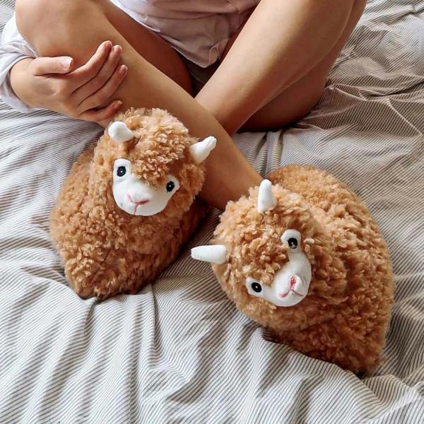 Close-up lifestyle view of a person wearing fluffy brown plush alpaca slippers while sitting on a striped bed sheet.