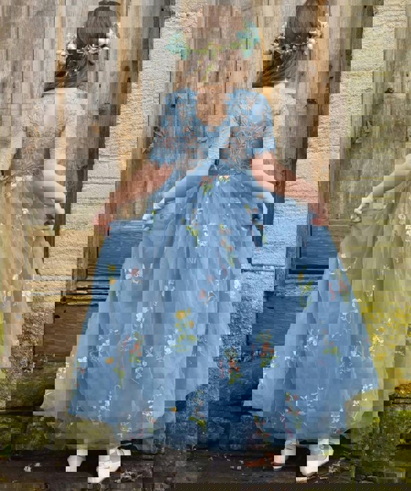 Young girl in a blue floral dress standing against a rustic wooden and stone wall.