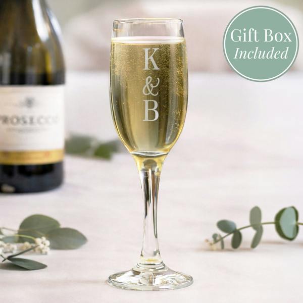 An engraved champagne flute by Abode Home & Gifts filled with golden prosecco, standing on a light table next to eucalyptus sprigs and a blurred prosecco bottle. The glass is custom engraved with the letters "K", an ampersand, and a "B" stacked vertically. A green graphic circle in the corner reads "Gift Box Included".