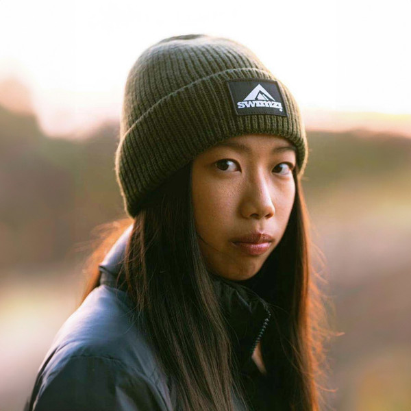 Young woman wearing a simple beanie hat knitted in a chunky single rib in a muted shade of green. There is a SWIMZI badge sewn to the front of the deep turn back headband.