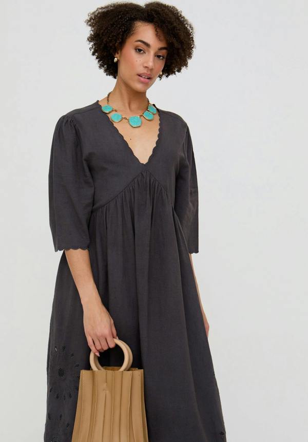 Woman wearing a dark gray dress with a V-neckline, holding a beige bag against a light gray background.
