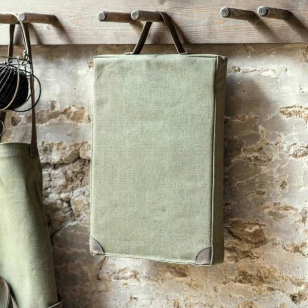 Khaki green canvas garden kneeler with brown faux suede handle hanging on a rustic wooden peg rack.