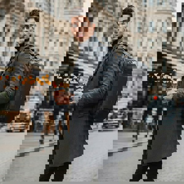 Man in a gray coat with a black backpack holding a coffee cup on a city street.