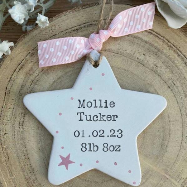 A hand-stamped white ceramic star personalised for Mollie Tucker hanging on pink polka dot ribbon over a wooden log slice.