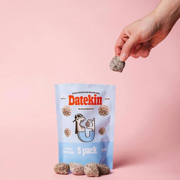 Hand holding a Datekin coconut snack ball with packaging on a pink background