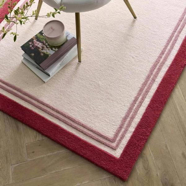 White rug with red border on wooden floor, books, and plant in the corner.