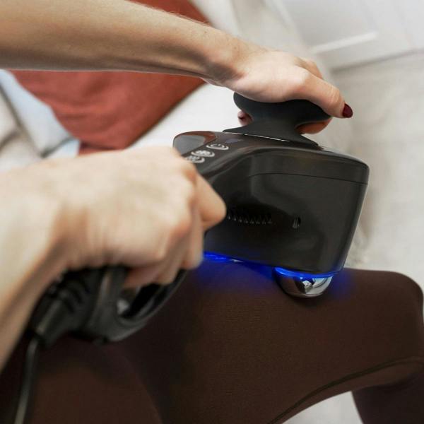 Indoors, someone uses the black Reviber Hot and Cold Percussion Massager with a blue light on their thigh for muscle relief; part of a couch and pillow can be seen in the background.