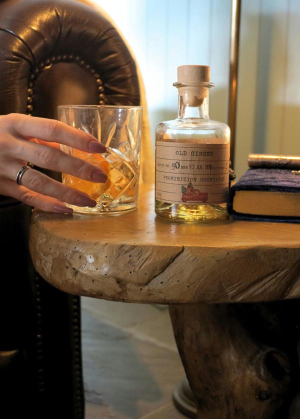 A hand holding a cut-glass tumbler filled with amber liquid beside a small clear bottle labeled 'OLD Ginger' on a rustic wooden table, with a leather chair and books in the background.