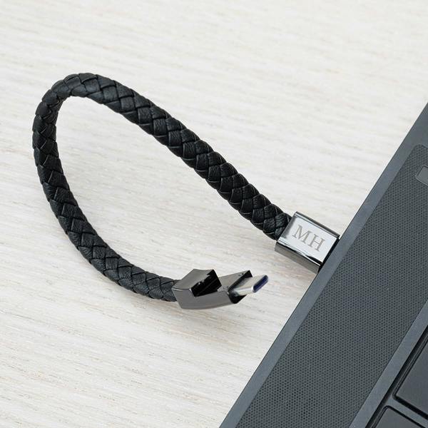 An unclasped braided black leather men's charging bracelet loop showing its gunmetal terminal hardware with the USB-C plug exposed near a laptop keyboard.