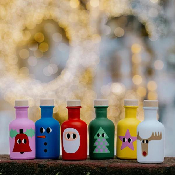 Six colourful miniature Christmas olive oil bottles standing in a row on a rustic ledge with a warm, glowing golden fairy light background.