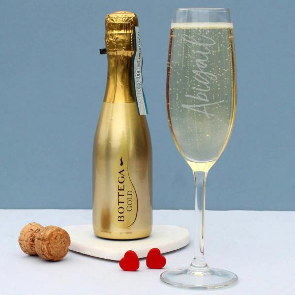 A miniature gold bottle of Bottega Gold prosecco next to a filled champagne flute engraved vertically with 'Abigail' on a white marble heart coaster.