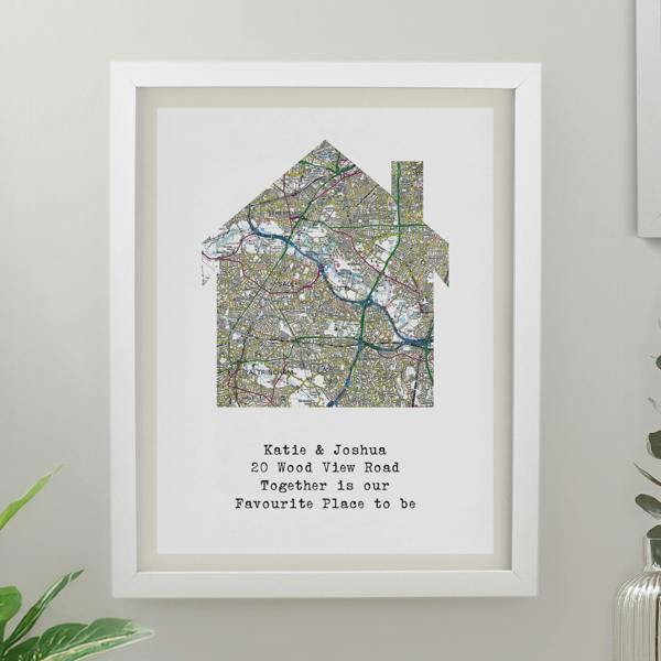 A white framed print showcasing a colourful Ordnance Survey map cut into a house silhouette, personalised with a typed three-line quote, sitting on a neutral wall.