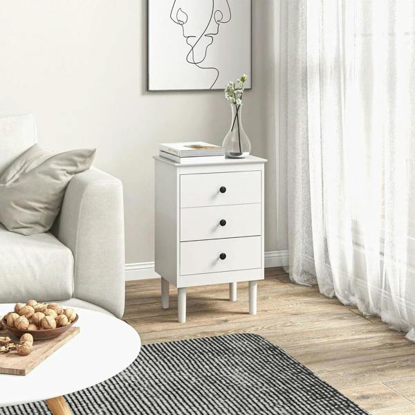White bedside cabinet with three drawers and round black knobs beside sofa, small vase.