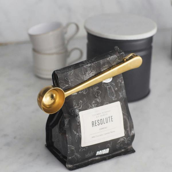 A round gold-coloured brass coffee scoop with a long clip handle attached across the top of a black coffee bean bag to seal it, styled on a marble counter.