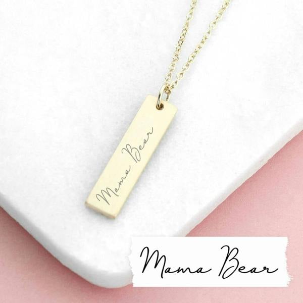 A matte gold vertical bar necklace reading Mama Bear in custom handwriting engraving, resting on a white marble edge with a pink background.