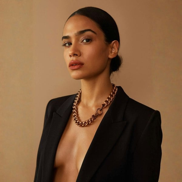 Woman wearing a black blazer and gold antique brass chain necklace against a beige background.