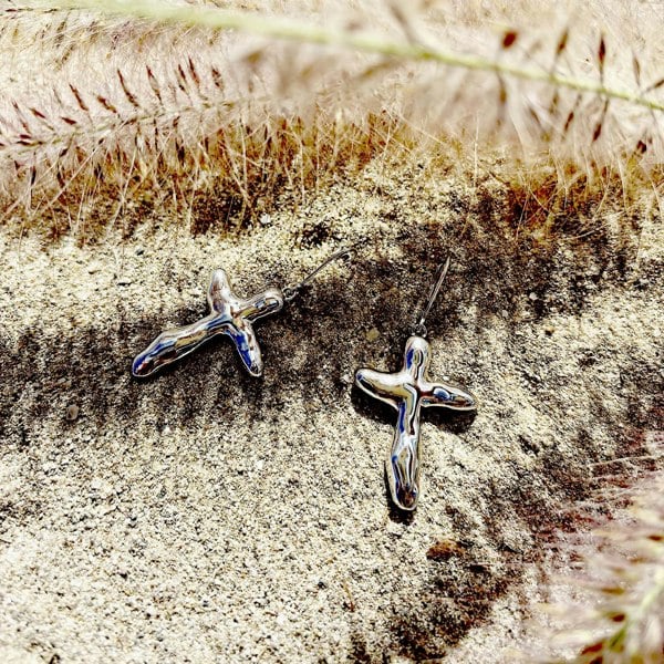 Silver cross irregular earrings photographed on sand.