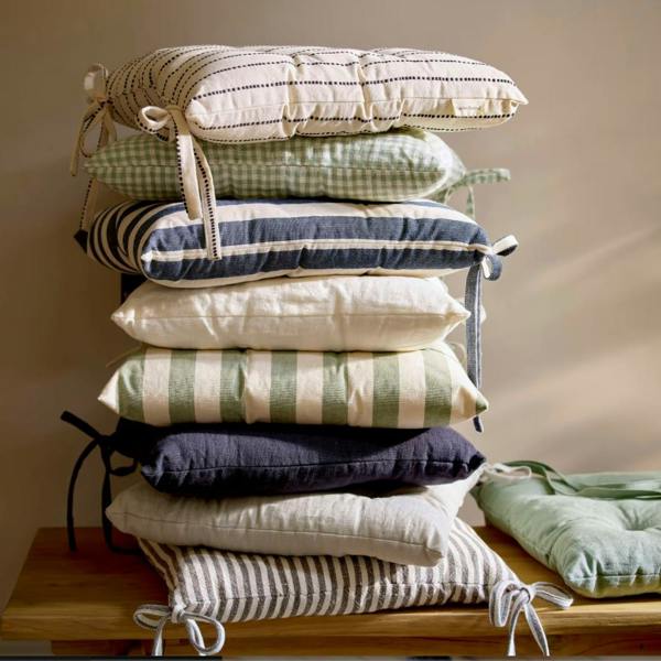 A stack of various linen seat pads, including Flint and Rosemary, on a wooden chair.