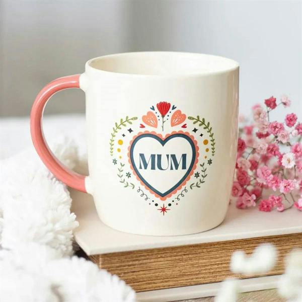White ceramic mug with coral-pink handle and “MUM” heart folk design