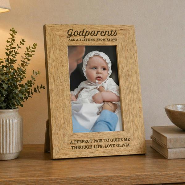 An engraved oak veneer photo frame displaying a baby in a white christening outfit, styled on a wooden tabletop with books and neutral home decor.