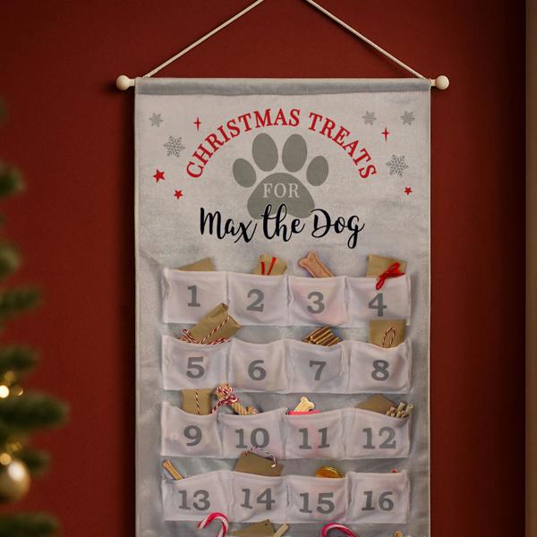 Alt Text: A personalised grey fabric advent calendar reading "Max the Dog" hanging on a dark red wall next to a blurred Christmas tree.