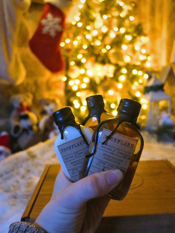 A hand holds three miniature decorative bottles labeled 'SNOWFLAKE' in front of a softly blurred Christmas tree with warm lights and a red stocking in the background.