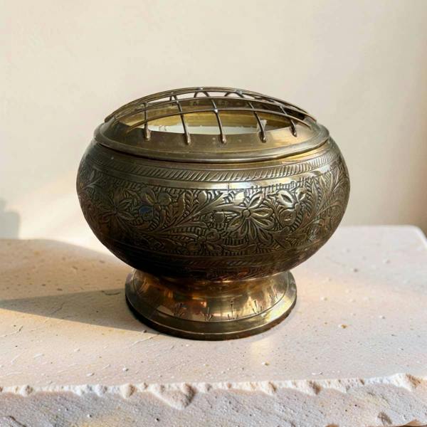 An ornate brass incense burner with intricate floral engravings and a wire lid, displayed on a light stone surface against a neutral background.