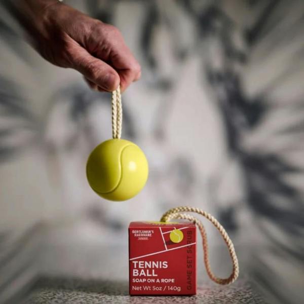 A hand holding up a yellow tennis ball shaped soap on a woven rope next to its red court-themed gift packaging.