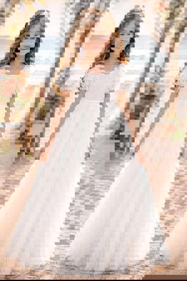 Young girl in a white lace dress standing on a beach with floral decorations.