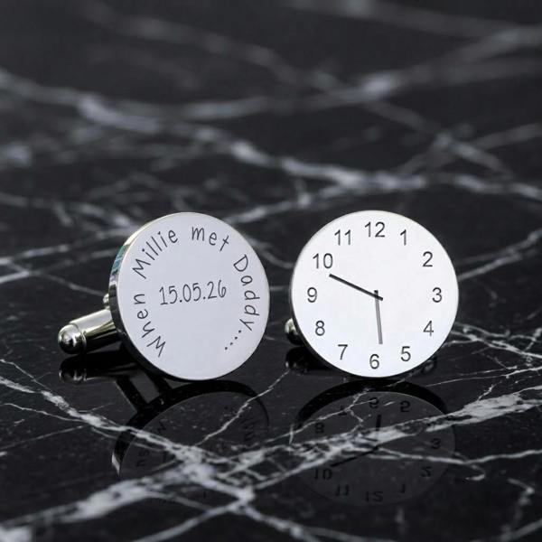 A pair of round silver rhodium-plated cufflinks displayed on a reflective black marble surface, one showing a birth clock face and the other engraved with a custom baby name and date.