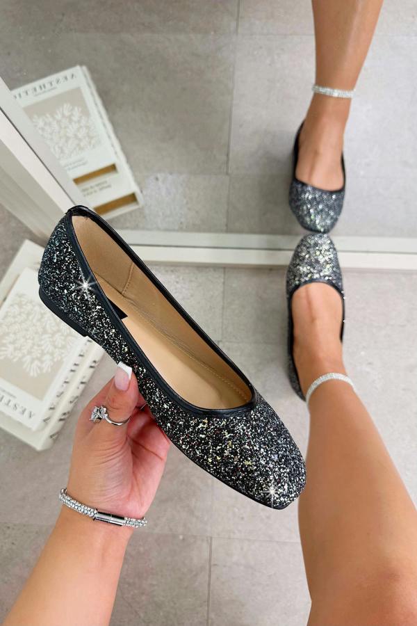 Black glittery flats being held up with a person wearing them on a tiled floor.