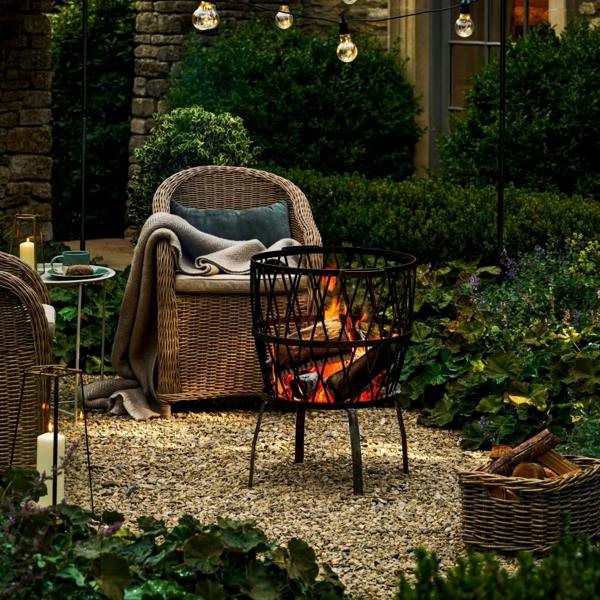 A Barrington Cross Fire Pit with a lit log fire, positioned on a gravel patio surrounded by wicker garden furniture and atmospheric festoon lighting.