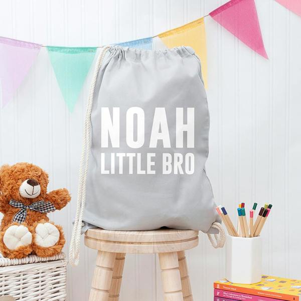 Boy's personalised light grey gym drawstring bag printed in white with NOAH LITTLE BRO, sitting on a wooden stool.