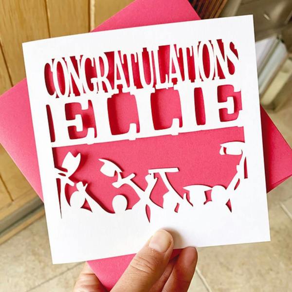 Hand holding a white papercut graduation card with Ellie cut out and pink background insert