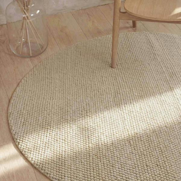 Round beige rug on a wooden floor with a glass jar and wooden chair.