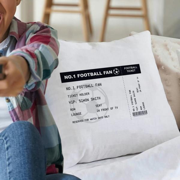A white polyester cushion printed with a custom black-and-white football ticket design showing the name 'Simon Smith', resting on a sofa next to a boy in a plaid shirt holding a TV remote.