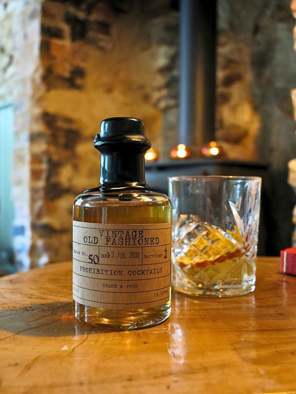 A small glass bottle of 'Vintage Old Fashioned' Prohibition Cocktail mix with a cream label and black cap, sitting on a wooden table next to a clear rocks glass with ice, set against a rustic stone wall and wood-burning stove background.