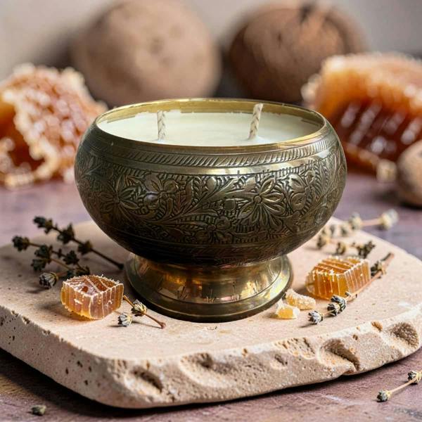 An ornate brass candle holder with floral engravings, filled with a white candle, styled on a stone slab with honeycomb, dried herbs, and walnuts in a warm, rustic setting.