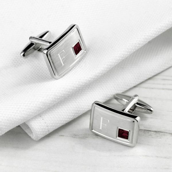 A pair of rectangular, rhodium-plated silver cufflinks resting on a white fabric surface, each featuring a polished silver face engraved with the letter 'F' and a square, deep red ruby-coloured crystal inset.