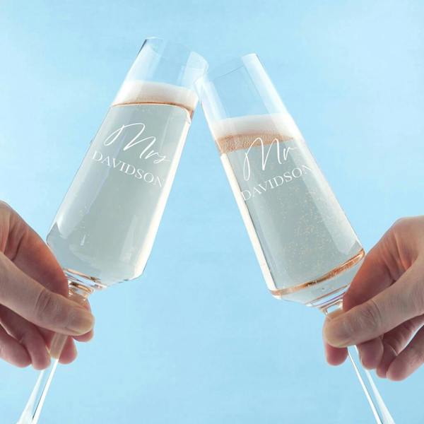 A close-up shot of two hands clinking two personalised champagne flutes filled with sparkling wine in a toast, featuring white engraved text displaying Mrs Davidson and Mr Davidson against a soft blue background.
