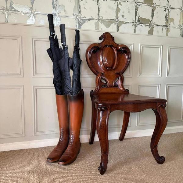 A faux-leather riding boot umbrella stand packed with three black umbrellas, positioned next to an ornate carved dark wood antique chair against a panelled hallway wall.