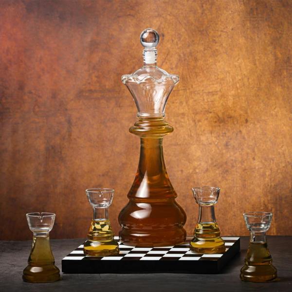 A striking glass chess decanter shaped like a queen sits on a black and white wooden chessboard base, flanked by four smaller, rook-shaped shot glasses filled with amber liquid. The set is captured against a warm, textured bronze background, emphasizing the elegant glass craft.