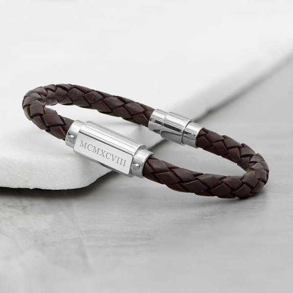 A close-up shot of the Personalised Men's Roman Numerals Woven Leather Bracelet in brown, resting on a white stone tile, displaying engraved Roman numerals on the stainless steel plaque.
