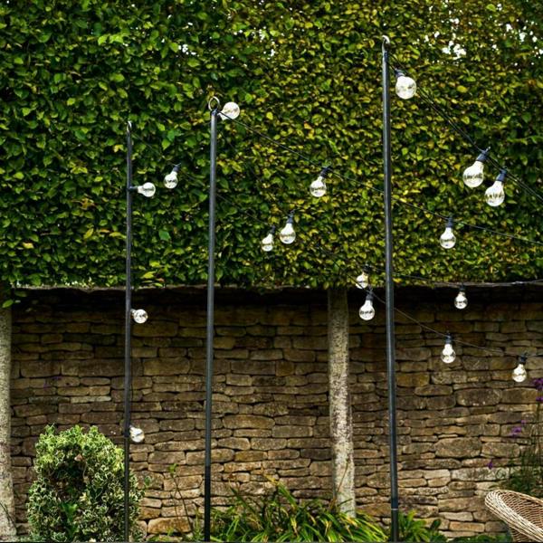 Three industrial raw metal festoon lighting poles supporting strings of warm white bulbs in front of a stone wall and high hedge.