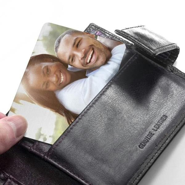 A hand pulling a custom metal wallet insert card featuring a printed colour photo of a smiling couple out of a black leather wallet slot.