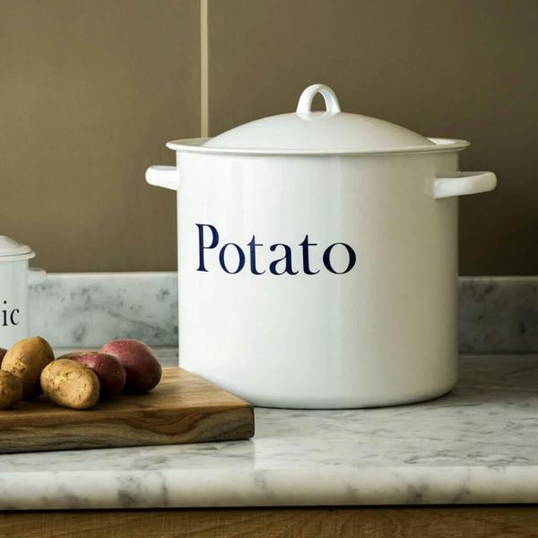 White enamel potato storage bin with navy Potato lettering resting on a marble kitchen countertop beside fresh potatoes.