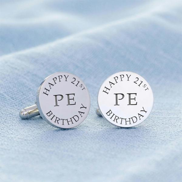 Pair of personalised silver round cufflinks engraved with "Happy 21st Birthday" and initials "PE" resting on a textured light blue tablecloth.