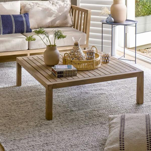 Square whitewash acacia wood coffee table styled with a tray and vase on a rug in a sunroom.